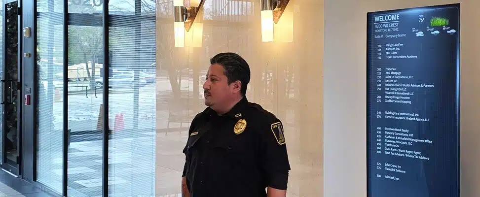 commercial-security-services-fortworth-img-1 Security guard in building lobby beside directory sign.